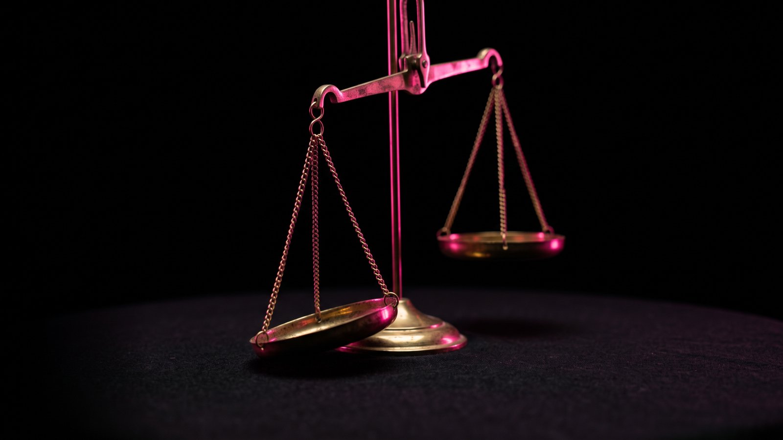 An antique brass balance scale with one pan tipped low under a single hot pink light against black, for an article on business email compromise