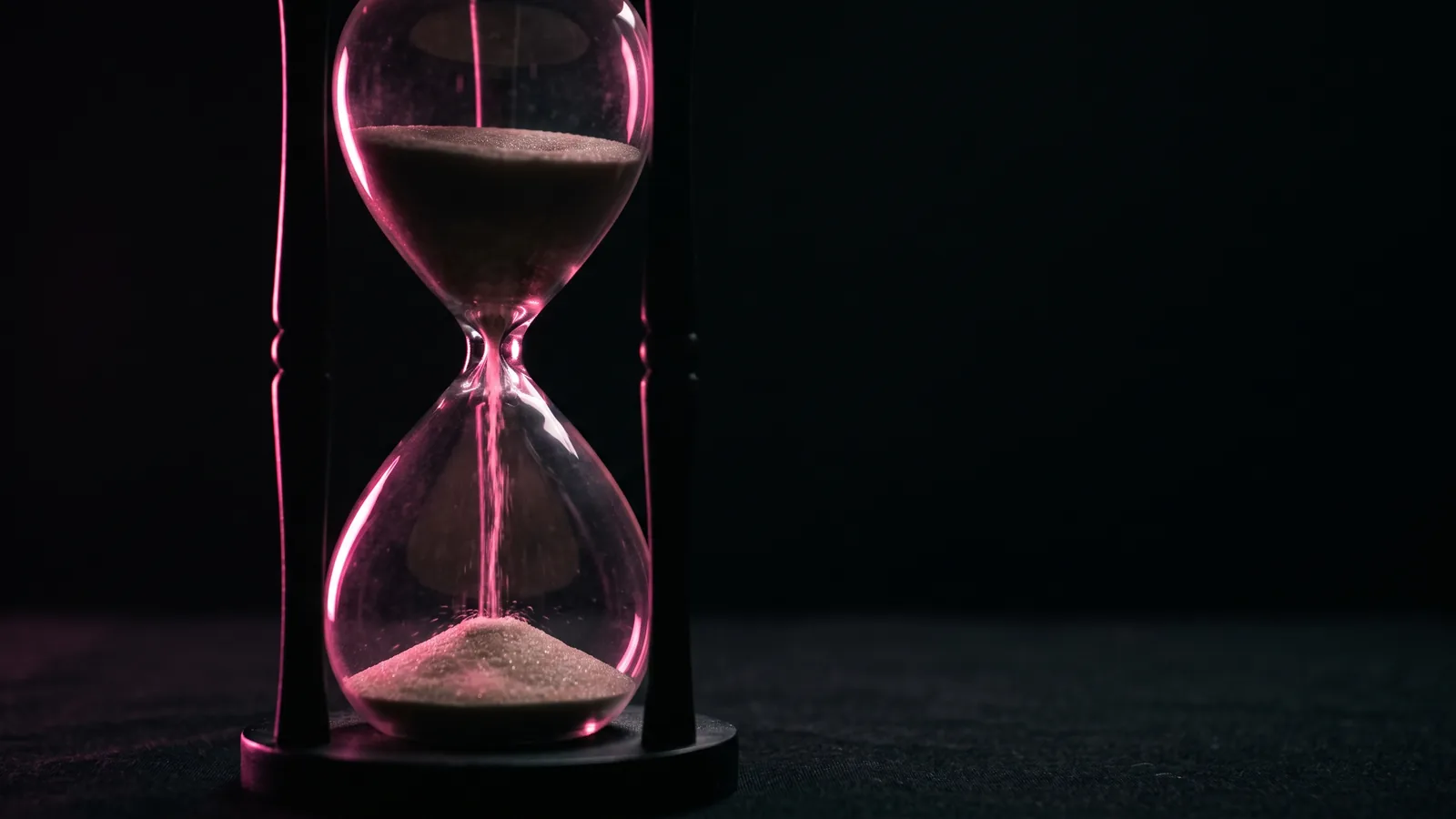 A glass hourglass on a black surface with sand falling through a beam of hot pink light, the first hour after a business email compromise wire