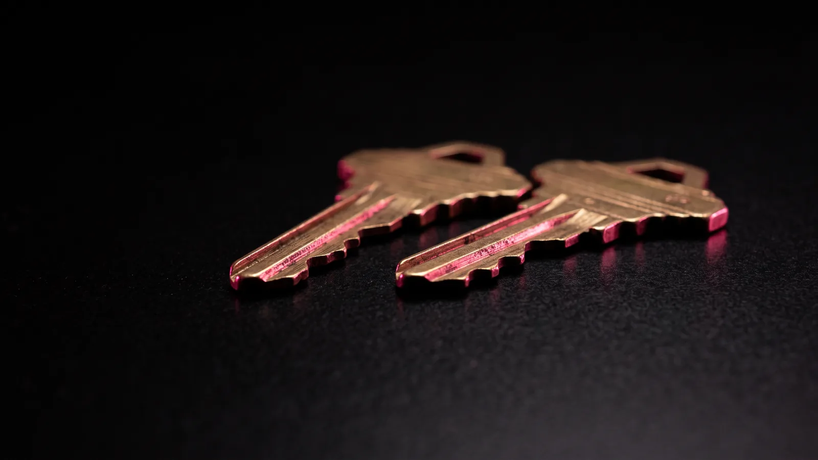 Two nearly identical brass keys side by side on black, one with a subtly different cut, under hot pink light, the lookalike domains used in business email compromise