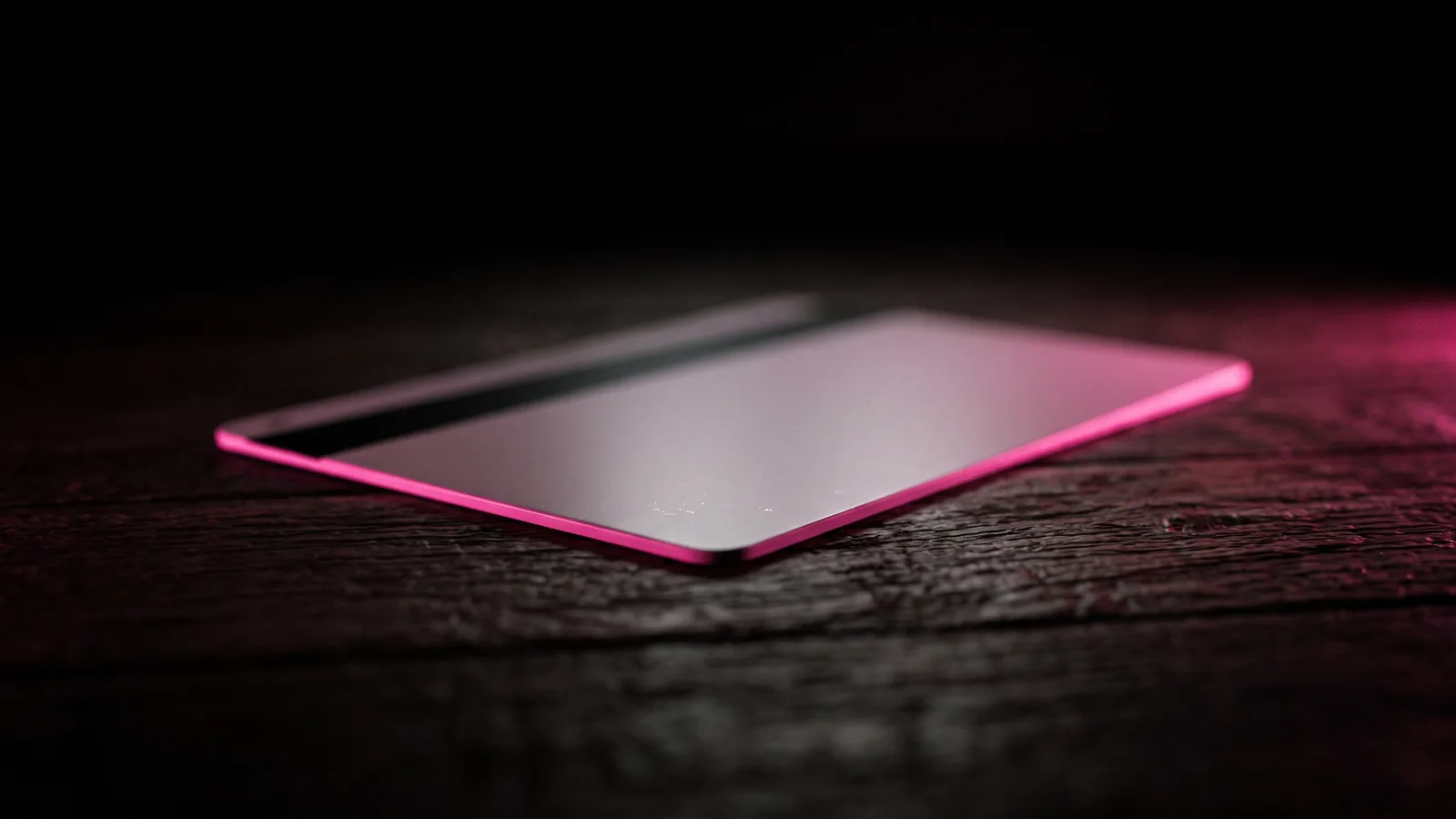 A blank hotel key card resting on dark wood with a thin hot pink edge light, an inherited breach that breach due diligence should surface before closing