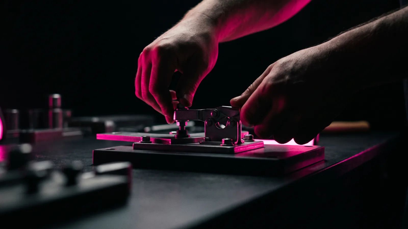 Two hands in shadow clamping a steel part into a workshop jig, the clamp edges lit hot pink against a black bench