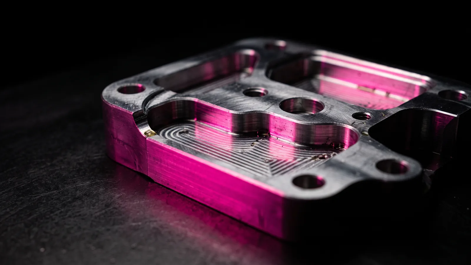 A machined aluminum part with fresh tool marks resting on black steel, one milled edge catching hot pink light