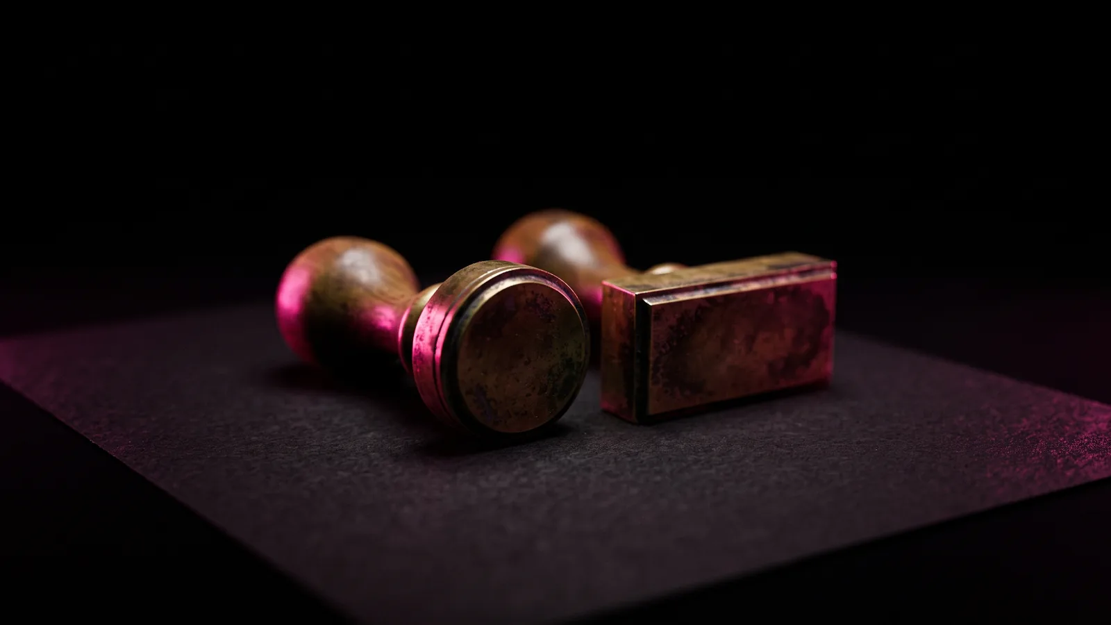 Two brass rubber stamps of different shapes standing side by side on black paper under hot pink light, comparing 201 CMR 17.00 and GDPR for Massachusetts companies