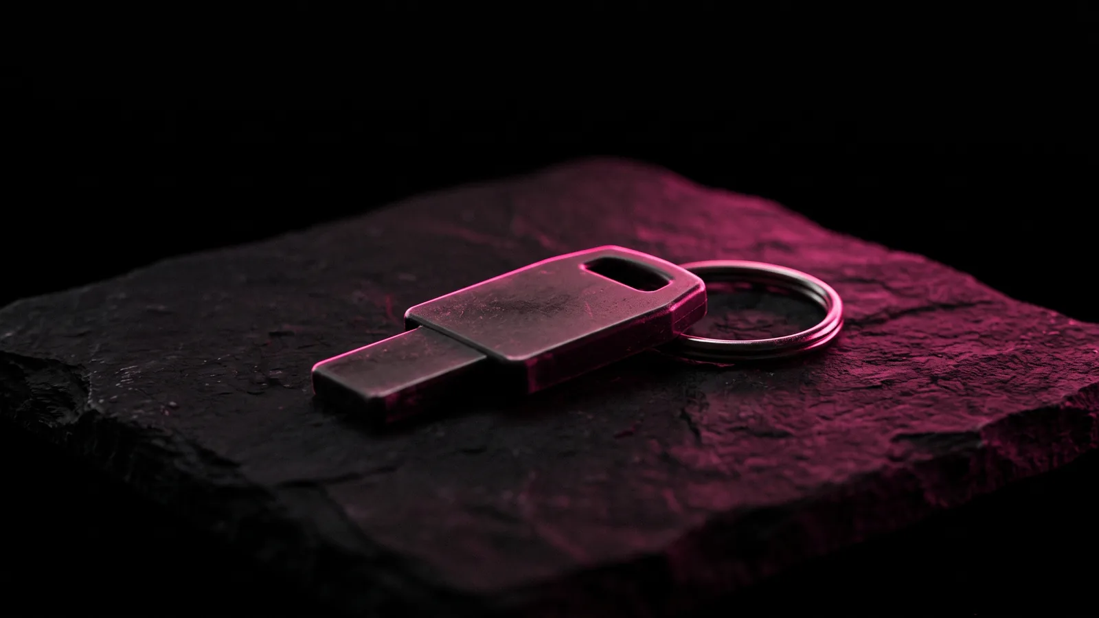A small hardware security key on a metal key ring resting on black stone, rim-lit in hot pink, the phishing-resistant answer to MFA fatigue