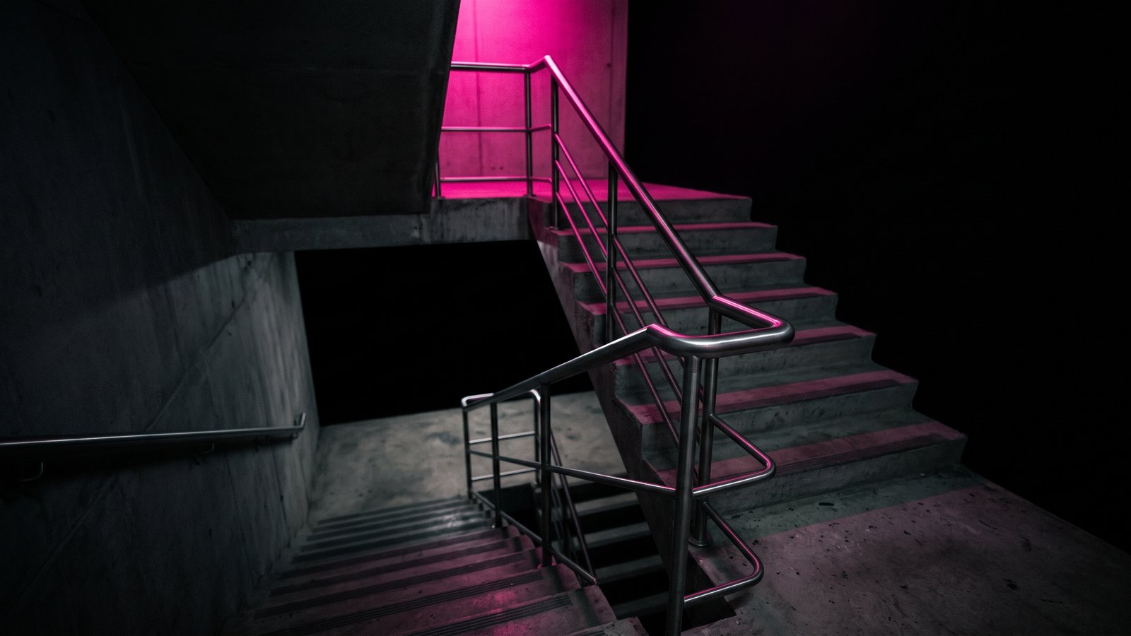 An empty concrete stairwell with its handrail traced in hot pink light against black, the phishing training cost of a drill