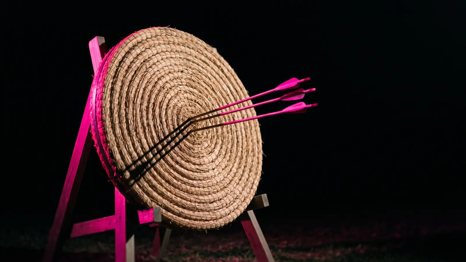 A straw archery practice target on a dark range with three arrows grouped just off center, rimmed in hot pink against black