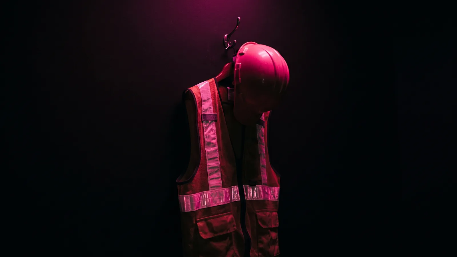 A high-visibility vest and a hard hat hanging on a single hook against black, lit only by hot pink light, the costume behind pretexting