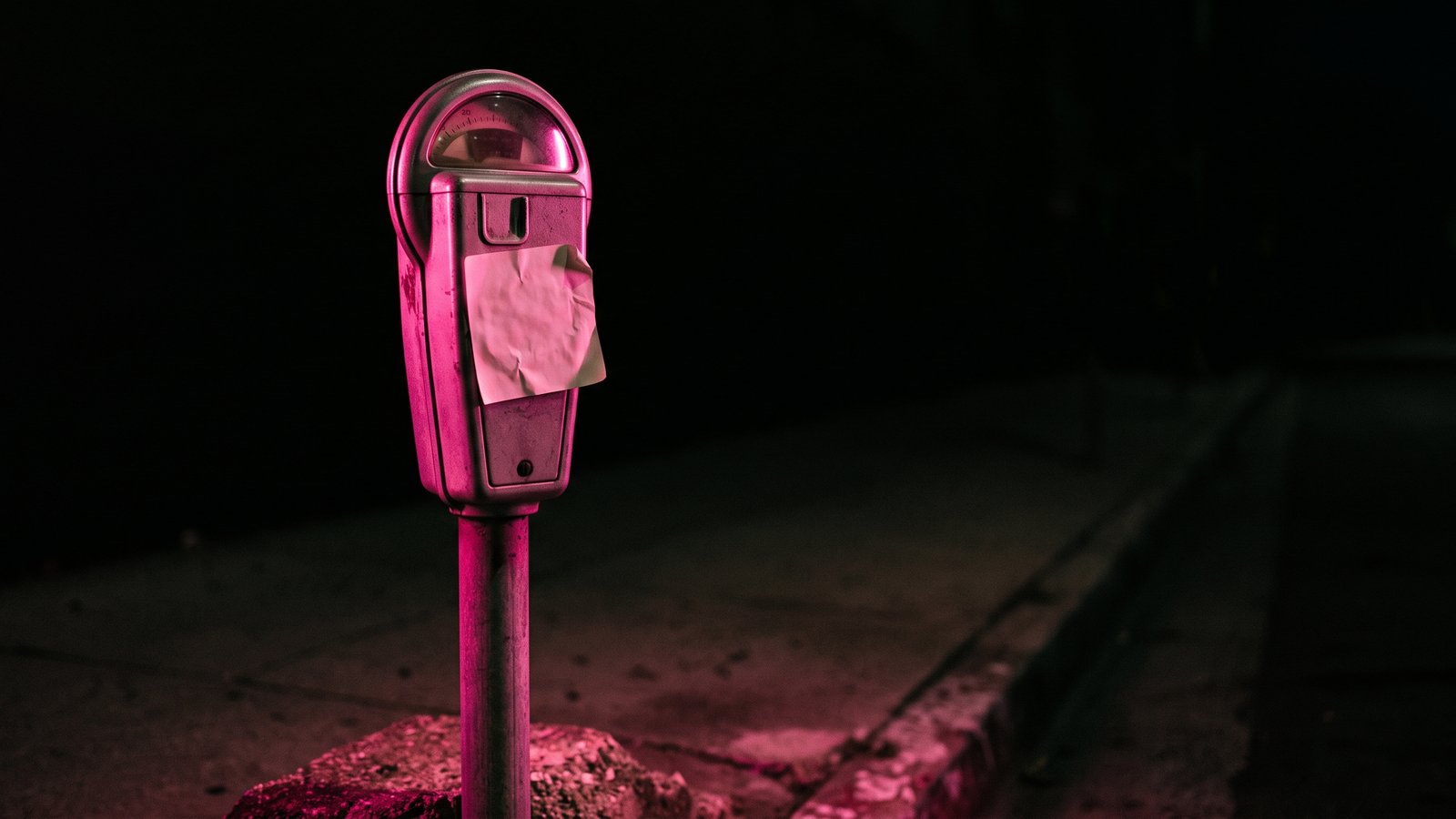 A street parking meter at night with a square sticker pasted over its panel, lit by hot pink light against black, for an article on what is quishing