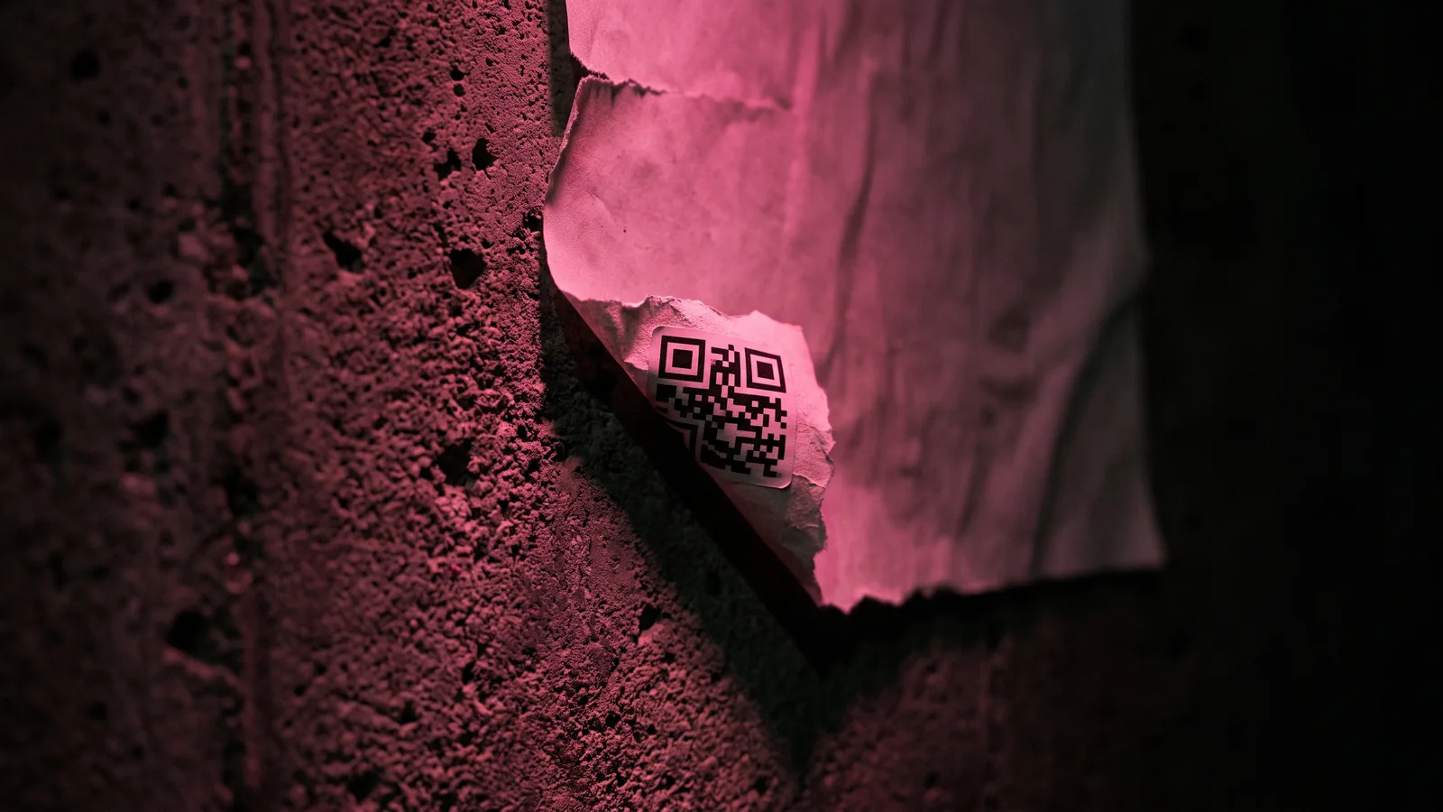 A torn poster on a bare concrete wall with a square black and white sticker layered over one corner, lit hot pink, showing how quishing codes cover real ones