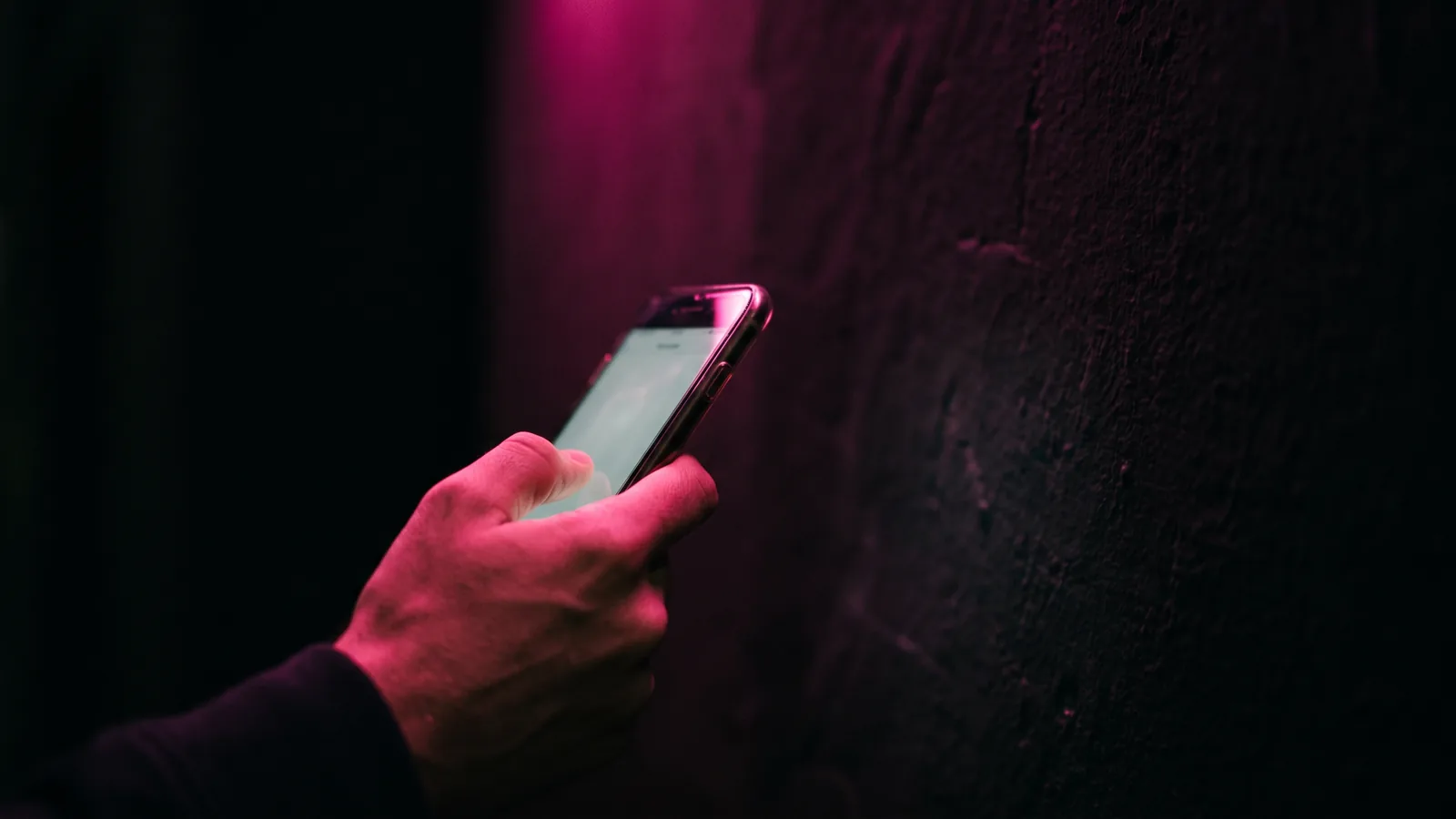 A hand holding a smartphone up toward a dark wall, the phone screen a soft unreadable glow, lit by hot pink light with shallow depth of field, the moment of a quishing scan