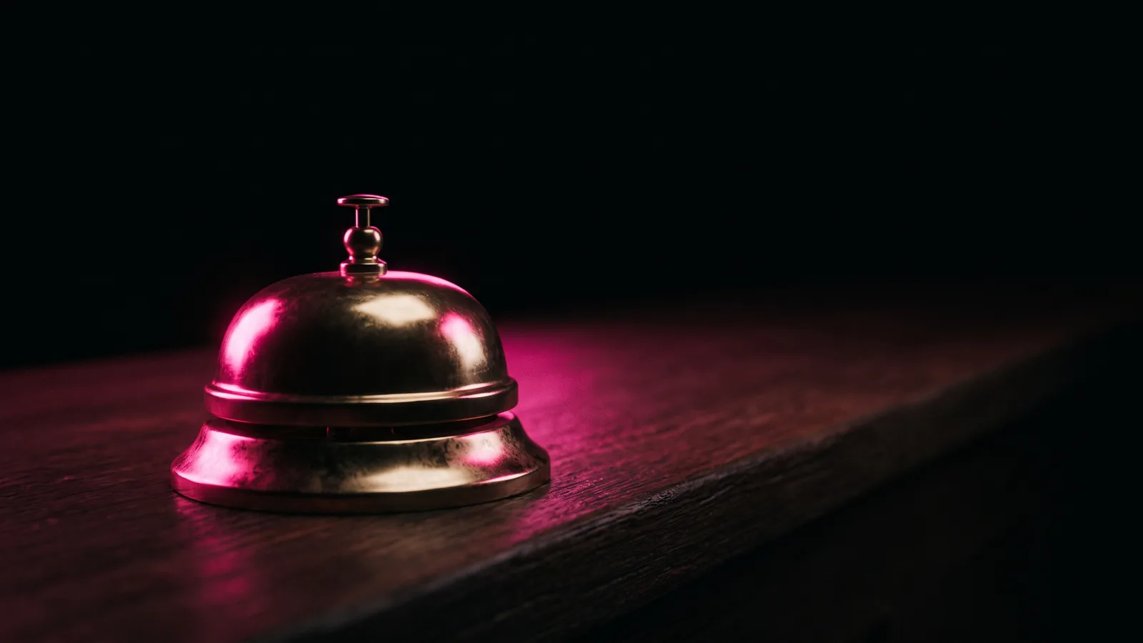 A brass service bell on a dark bank counter, its dome glinting hot pink against black, calling the bank first after you check if a message is a scam