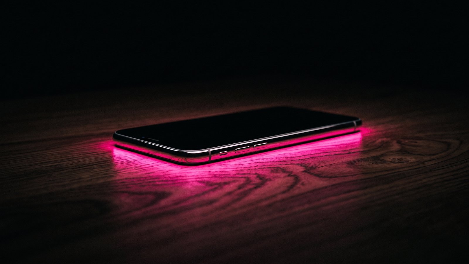 A smartphone lying face down on a dark wooden desk with hot pink light spilling from beneath its edges, for an article on what is smishing