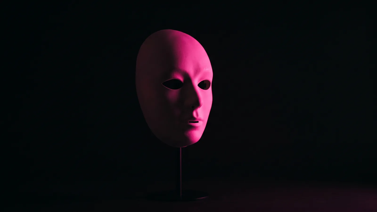 A plain white theatrical mask on a stand in darkness with hot pink light across one side, impersonation messages aimed at company social media accounts