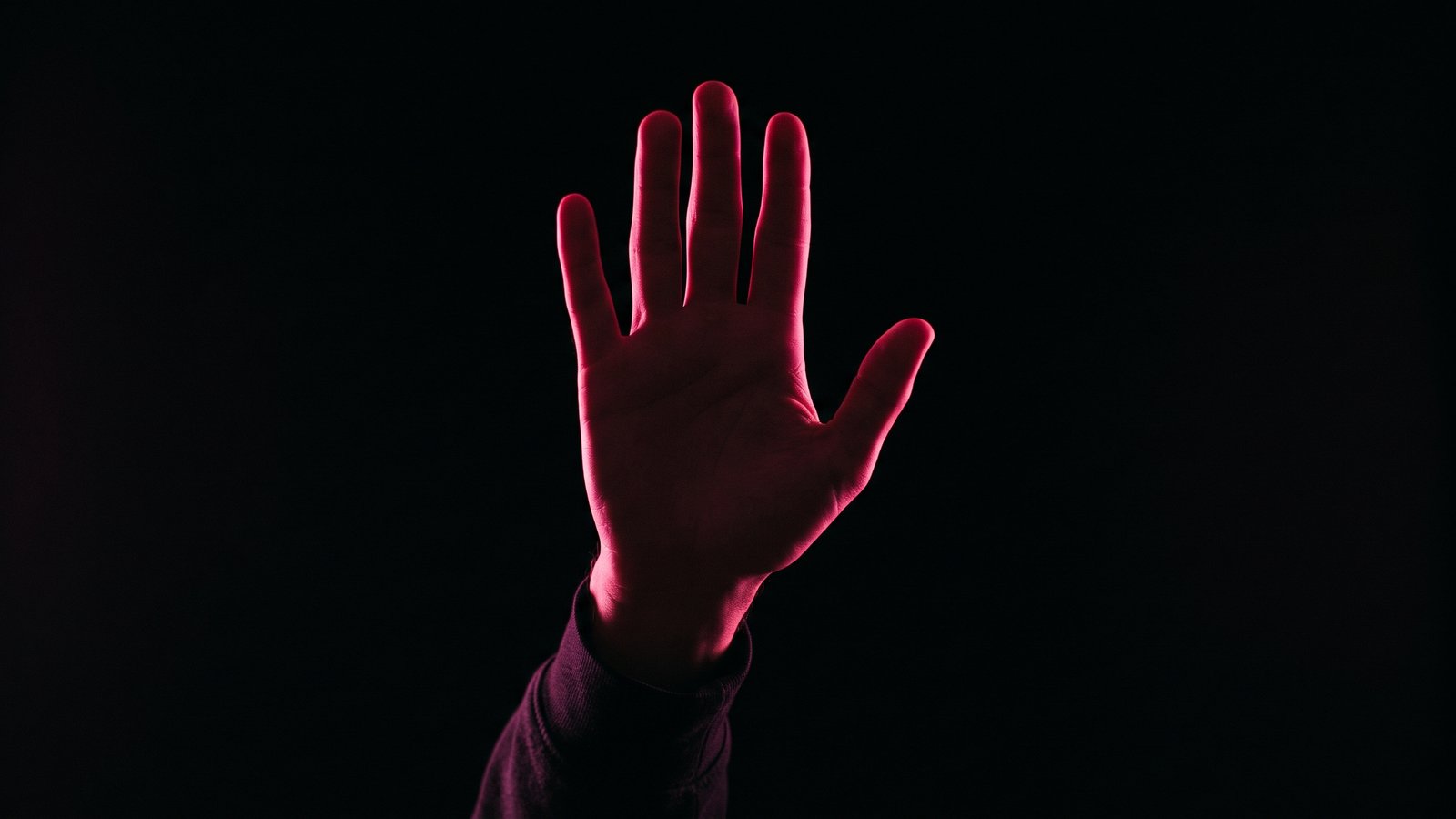 A raised open hand, palm toward the camera, in silhouette against black with hot pink rim light tracing the fingers, stopping before acting on a suspicious email