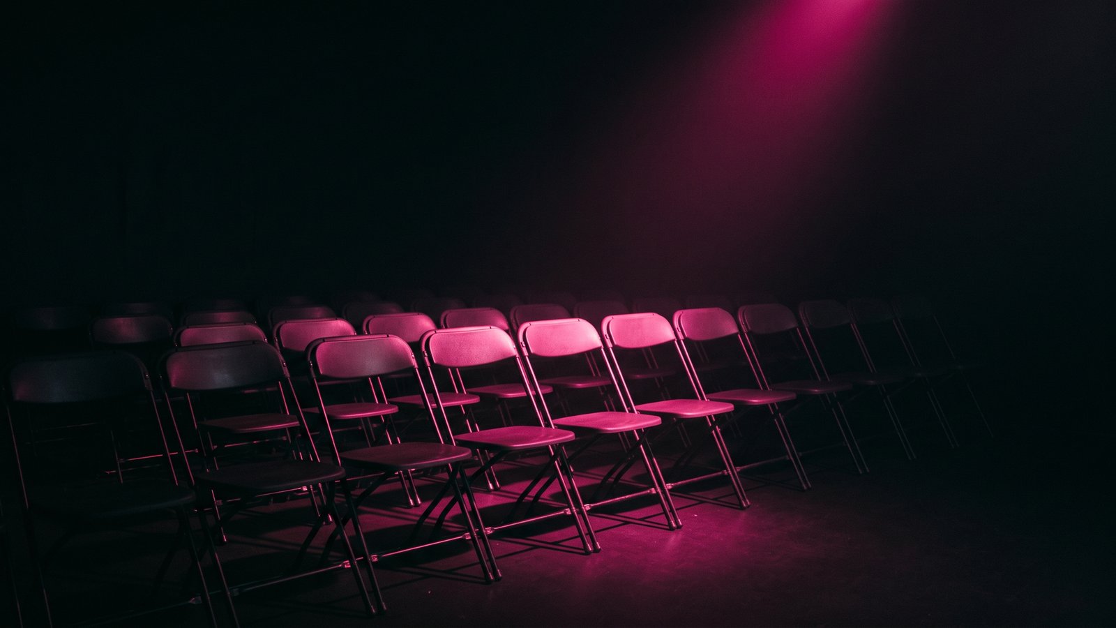 Rows of empty folding chairs in a dark training room with a single hot pink light falling across the front row, the gap between security awareness training and behavior